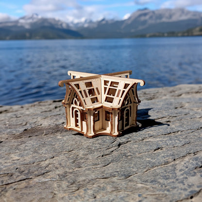 Small laser cut plywood architectural puzzle greenhouse on a riverside timber bench under a bright sky with soft clouds.
