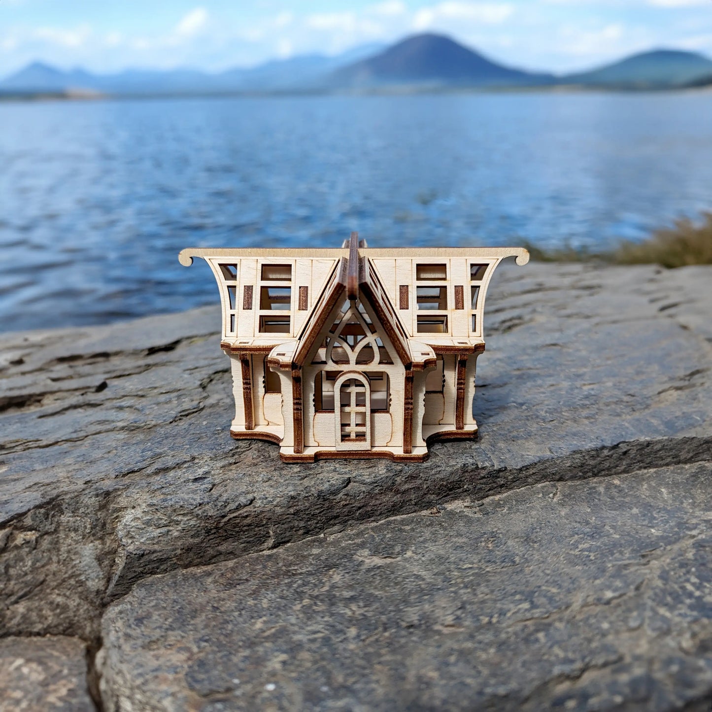 Straight front view of an intricate laser cut plywood glasshouse puzzle staged on rocks with a scenic lake and mountains.