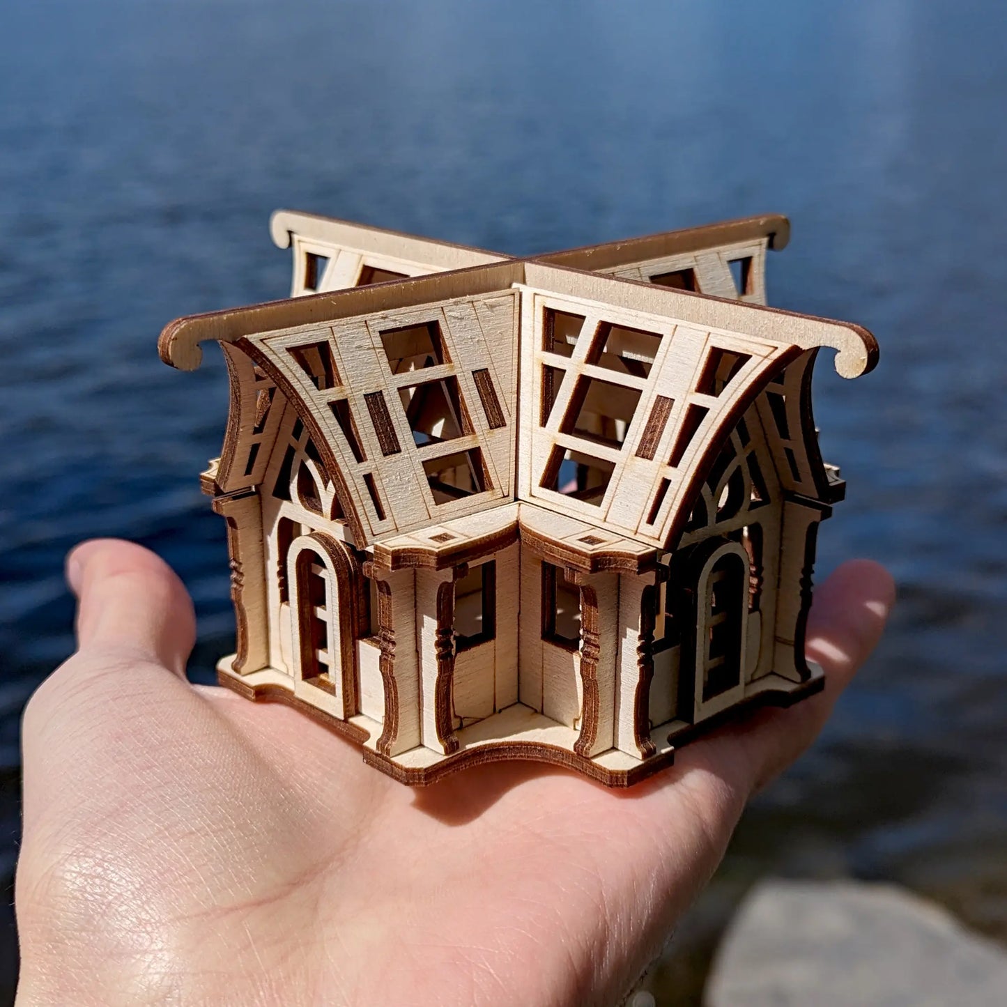 A small laser cut plywood winter garden pavilion model held gently in a hand against blurry ripple water backdrop.