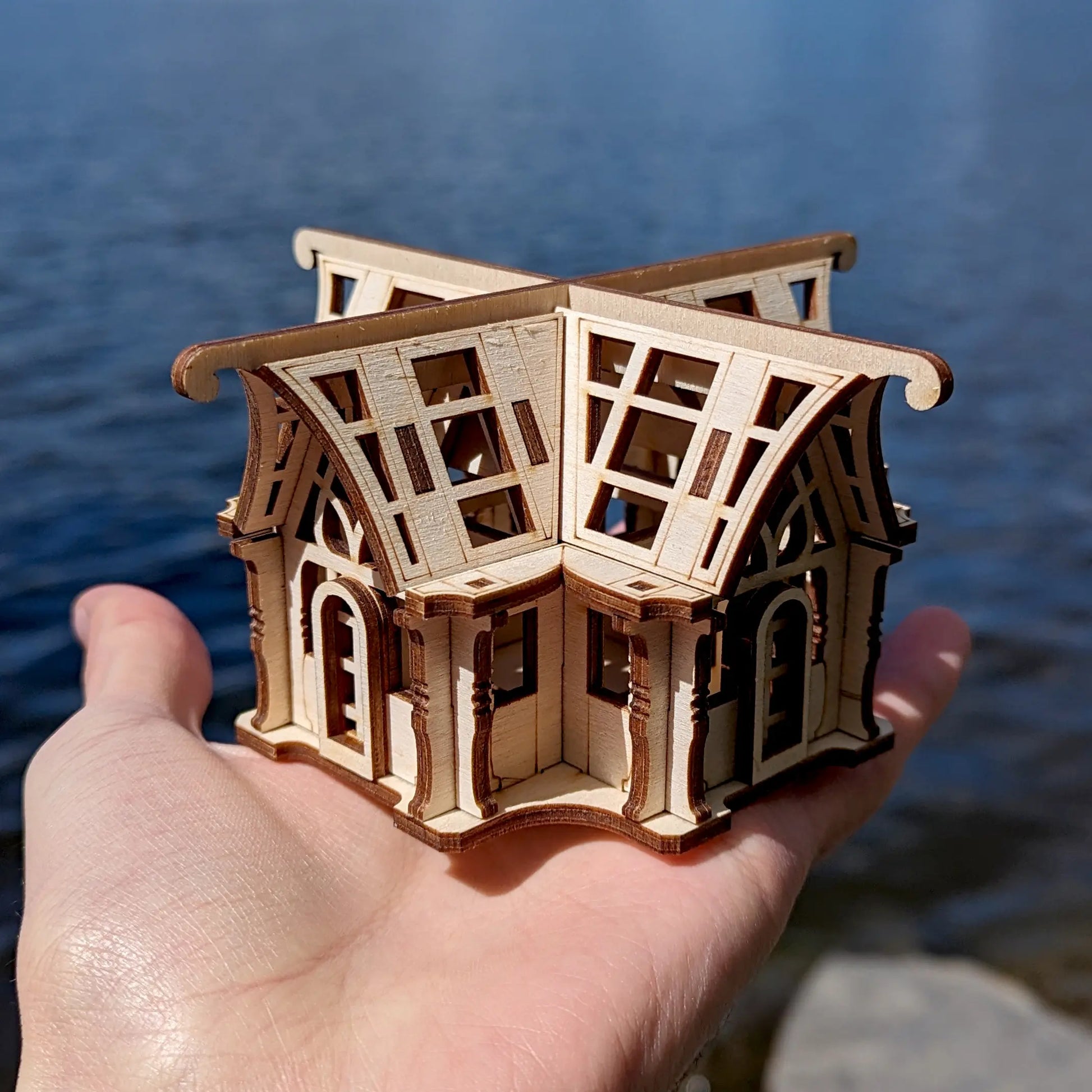 A small laser cut plywood winter garden pavilion model held gently in a hand against blurry ripple water backdrop.