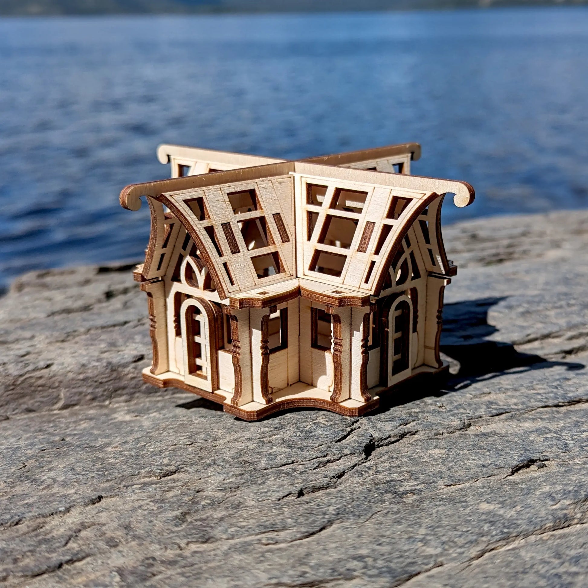 Miniature laser cut plywood greenhouse model on a rough stone surface with deep blue river water in the background.