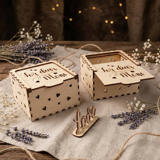 Two handcrafted laser-cut plywood gift boxes featuring "For dear Mom" engraved in elegant script on the sliding lids. The boxes have decorative heart-shaped cutouts on the sides and finger-joint corners. Styled in a rustic setting with bundles of lavender, dried flowers, and a small wooden "love" sign.