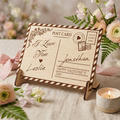 A personalized laser-cut wooden Valentine's Day postcard on a matching stand. Engraved with "I Love You", "Leslie", "Jonathan", and a vintage postmark. Features a striped border and floral corner design. Styled on a table with flowers and a candle.