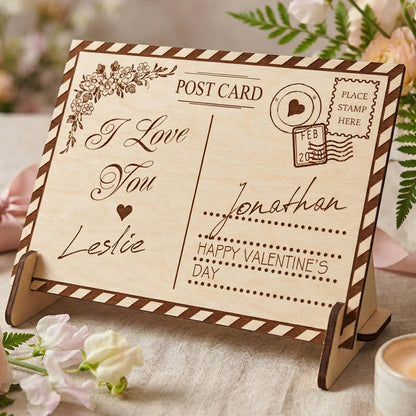 A personalized laser-cut wooden Valentine's Day postcard on a matching stand. Engraved with "I Love You", "Leslie", "Jonathan", and a vintage postmark. Features a striped border and floral corner design. Styled on a table with flowers and a candle.