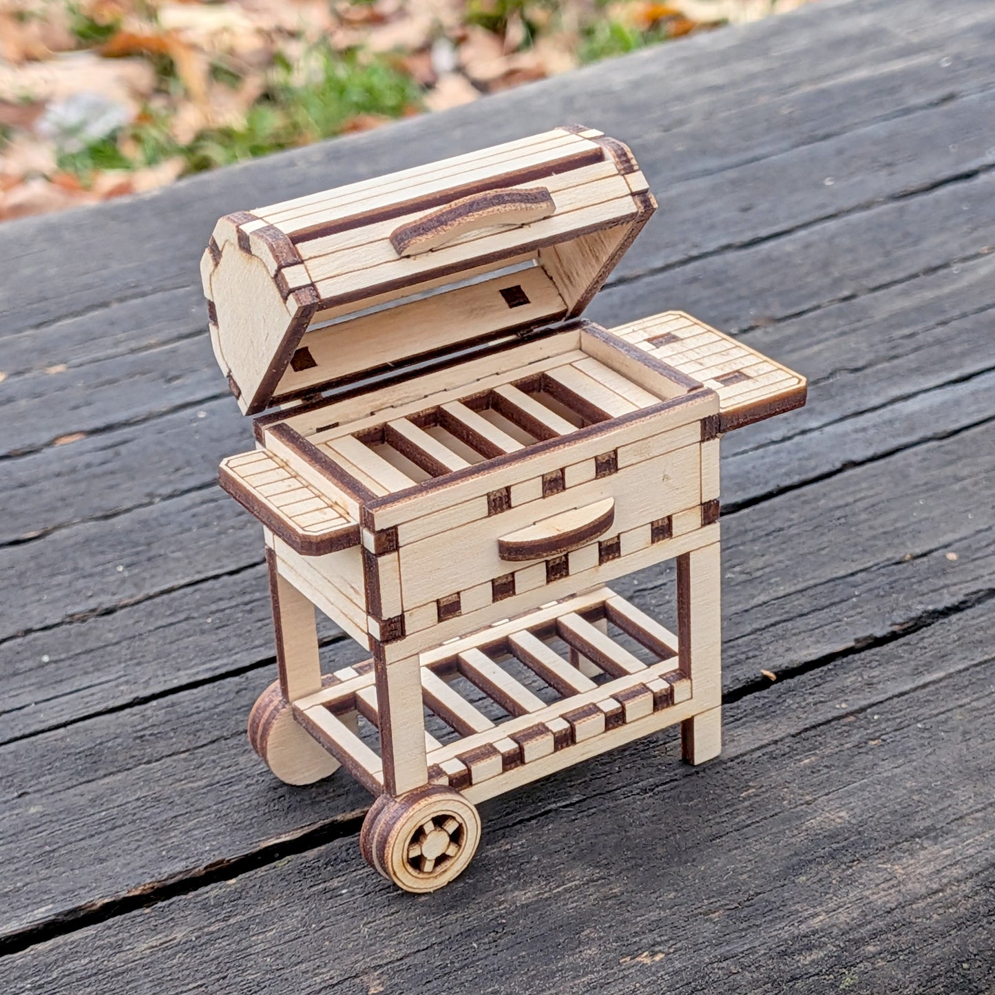 Laser cut miniature wooden BBQ grill model with an open lid, side shelves, and small wheels, displayed outdoors on a wooden surface.