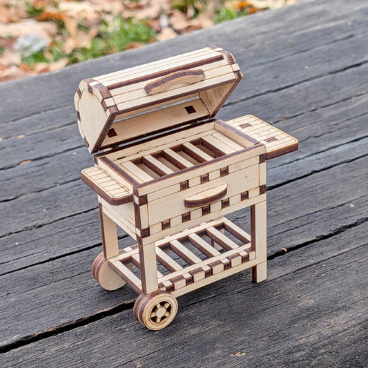 Laser cut miniature wooden BBQ grill model with an open lid, side shelves, and small wheels, displayed outdoors on a wooden surface.
