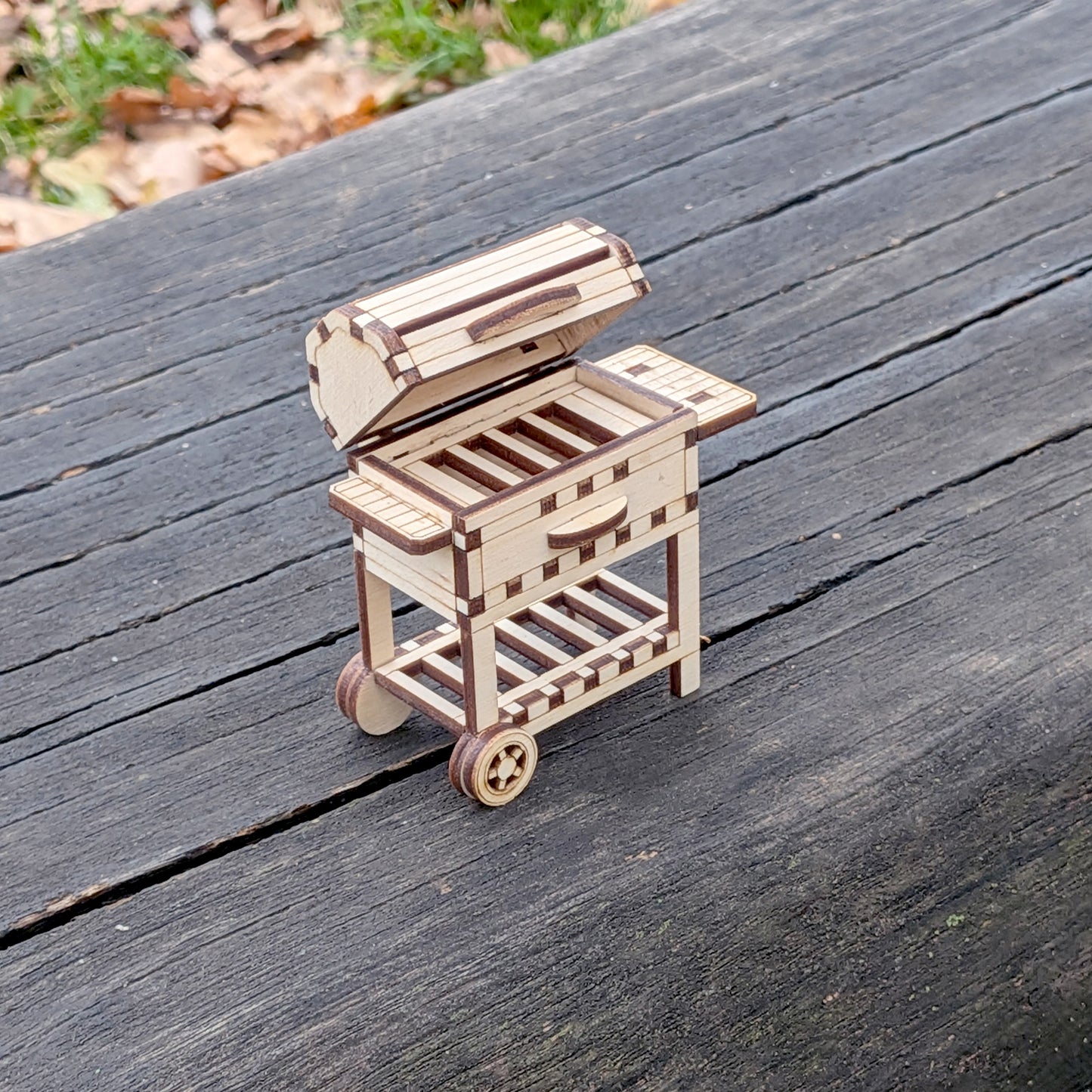 Miniature laser cut wooden BBQ grill viewed from above, placed on a weathered wooden table outdoors