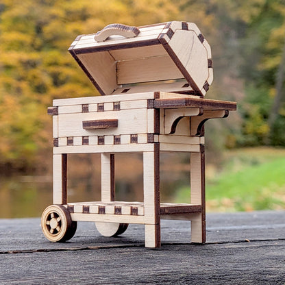 Side view of the miniature laser cut BBQ grill with open lid, showing the small wheels and bottom storage rack, photographed outdoors.