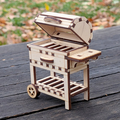 Laser cut wooden BBQ grill with lid open and side shelf visible, showing the detailed cut pieces and small wheels, photographed outside.