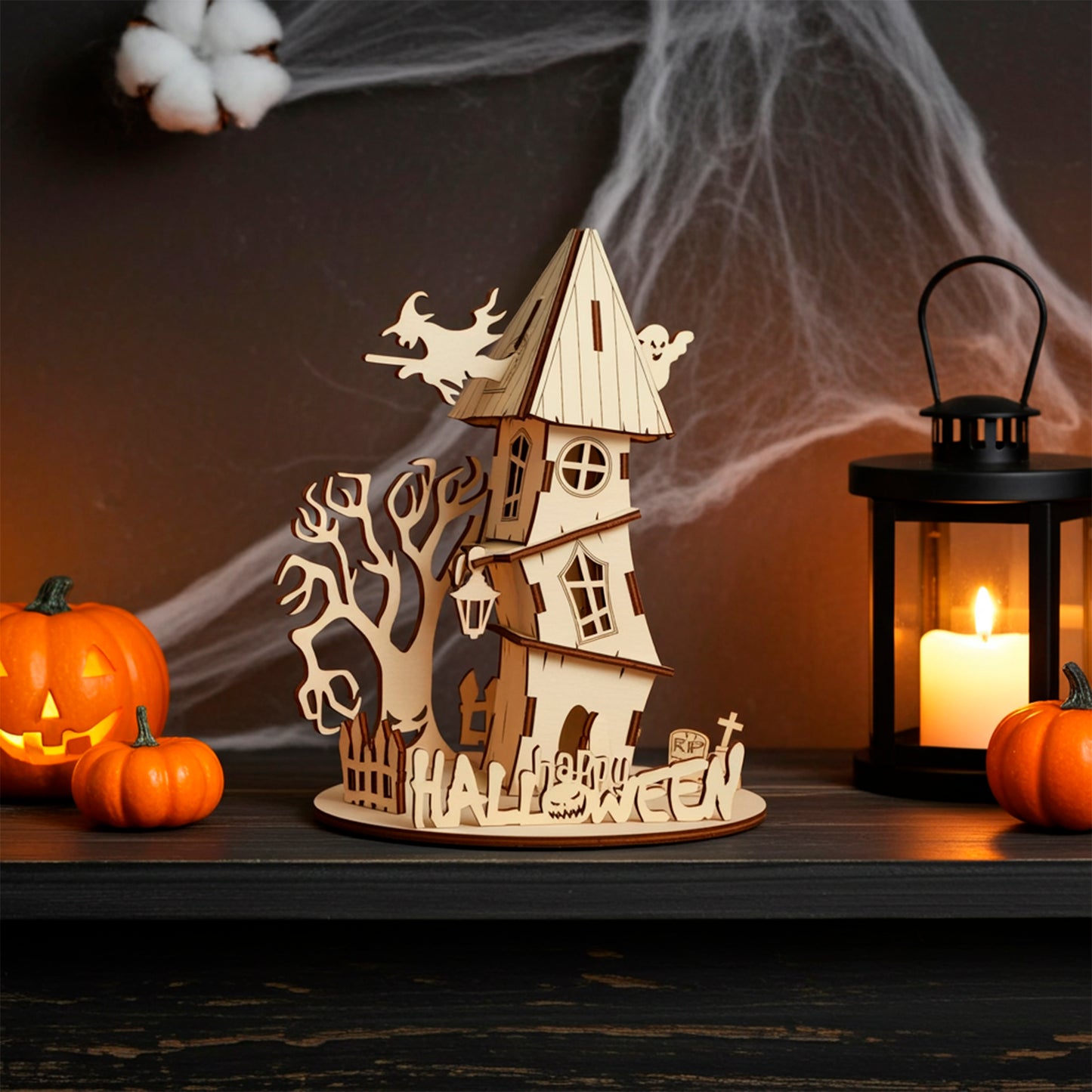 Laser cut plywood wooden haunted house decoration with a witch, ghost, spooky tree, and "Halloween" fence, set on a dark shelf with pumpkins, cotton web, and a candle lantern creating an eerie glow