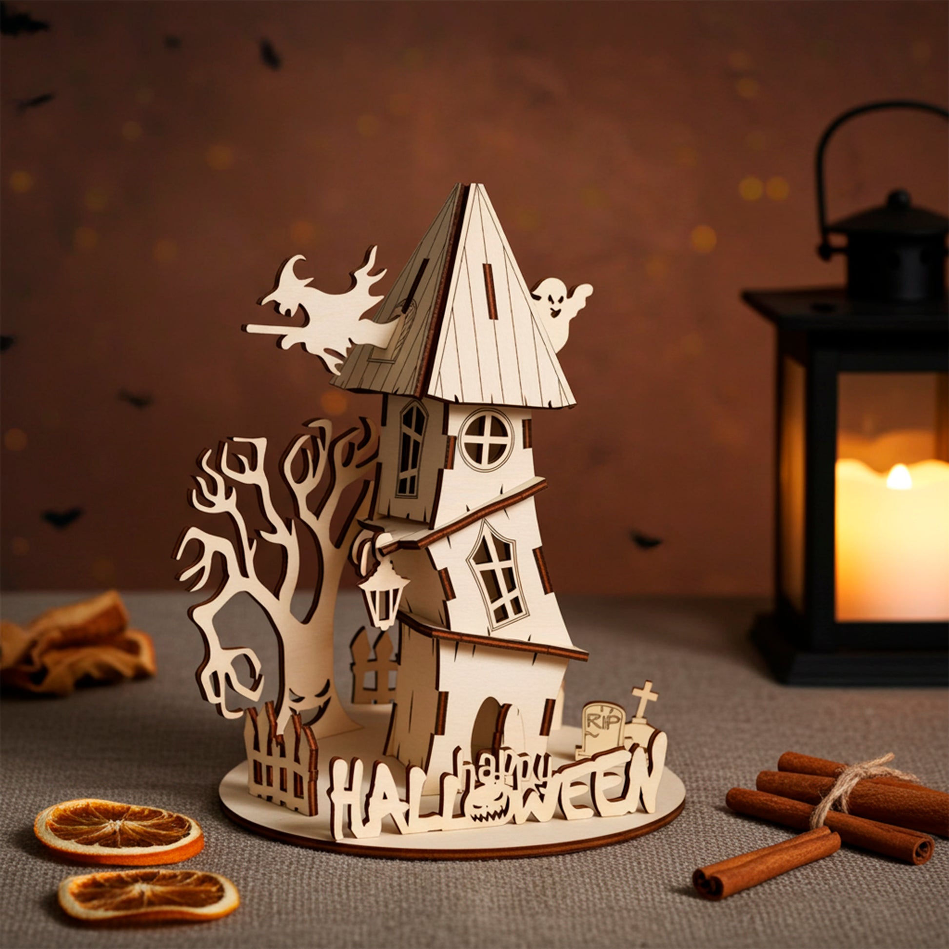 Laser cut plywood wooden haunted house decoration with a witch, ghost, spooky tree, and "Halloween" fence, set on a dark shelf with pumpkins, cotton web, and a candle lantern creating an eerie glow