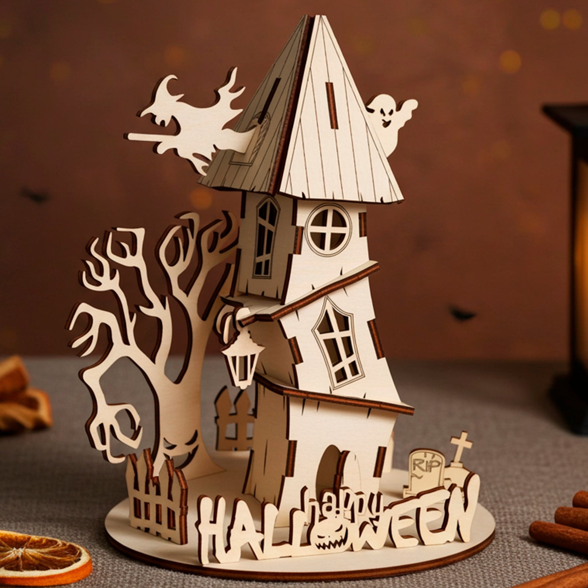 Laser cut plywood wooden haunted house decoration with a witch, ghost, spooky tree, and "Halloween" fence, set on a dark shelf with pumpkins, cotton web, and a candle lantern creating an eerie glow