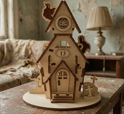 Front view of a two-story haunted house made from laser cut plywood. Features a "13" plaque, a jack-o'-lantern in the attic window, and ghost silhouettes. A miniature graveyard and spooky tree sit on the circular base.