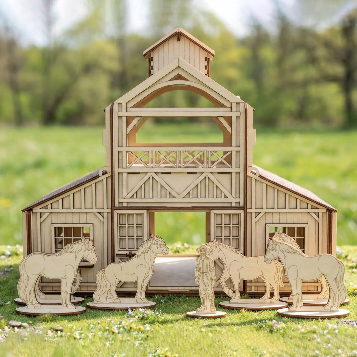 Straight front view of a laser cut plywood horse barn model placed outdoors on green grass with cutout toy figurines.