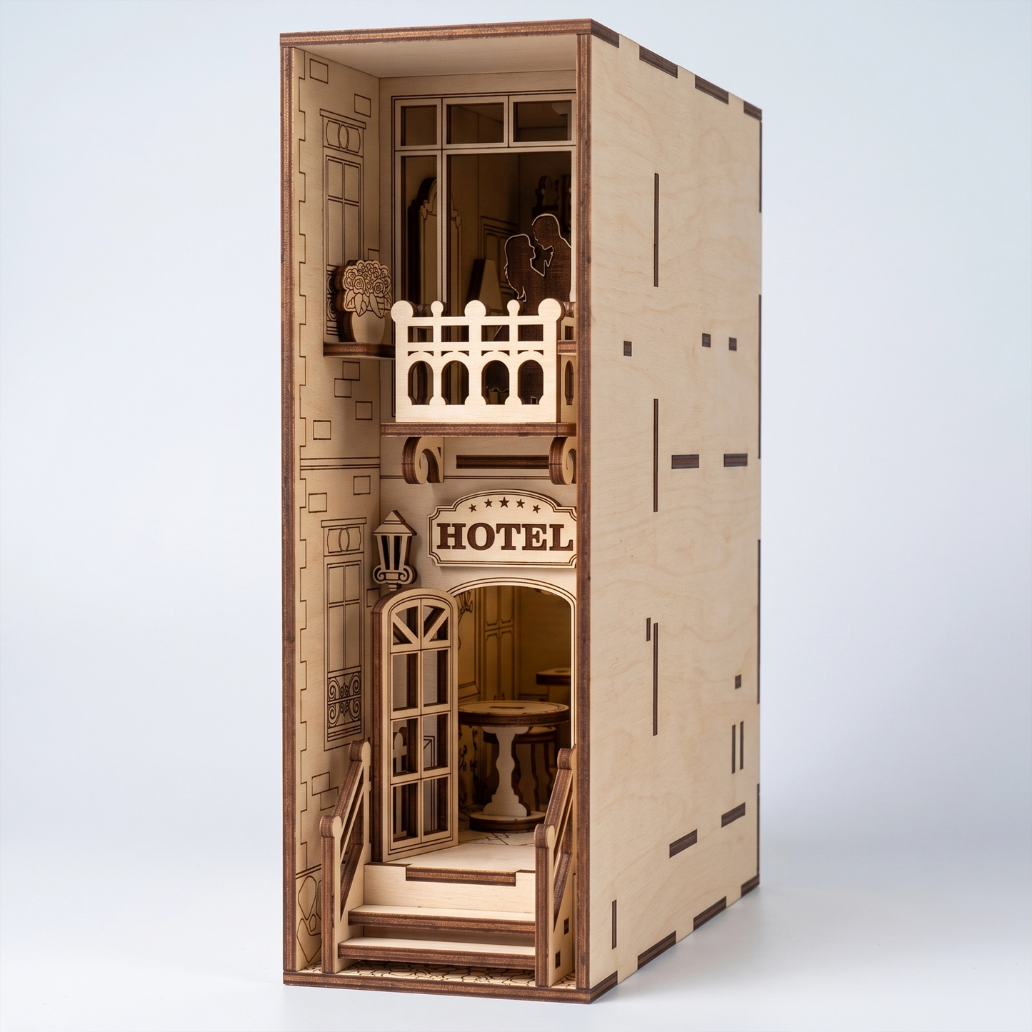 Three-quarter view of a light birch plywood laser-cut hotel book nook. Features a balcony with couple, entrance sign, and natural dark burnt edges.