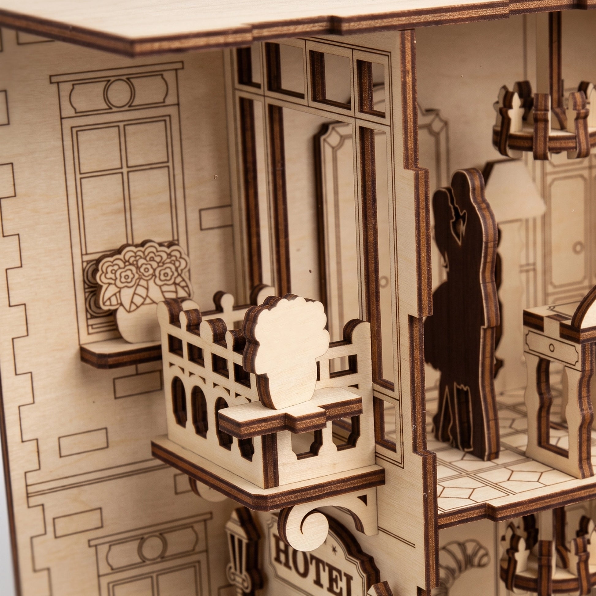Detailed view of laser-cut birch plywood hotel balcony with flower pot and silhouette couple. Natural light wood grain with dark, realistic burnt edges.
