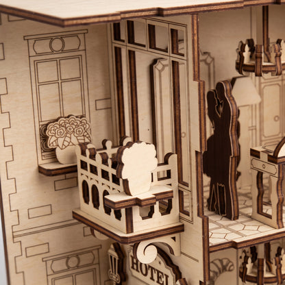 Detailed view of laser-cut birch plywood hotel balcony with flower pot and silhouette couple. Natural light wood grain with dark, realistic burnt edges.