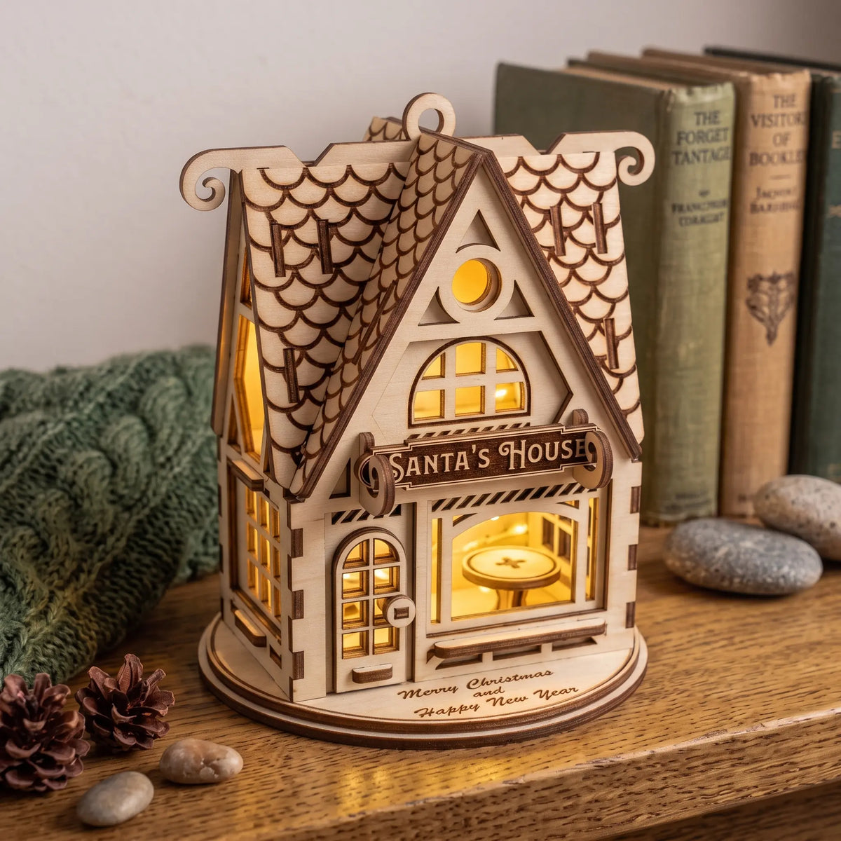 Lit laser cut plywood Santa House model resting on a wooden bookshelf by vintage books and pinecones.