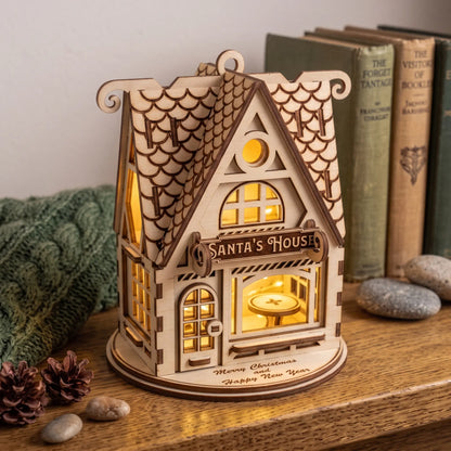 Lit laser cut plywood Santa House model resting on a wooden bookshelf by vintage books and pinecones.