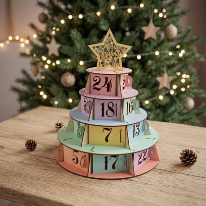 Pastel laser cut plywood Christmas tree advent calendar on wooden tabletop in front of decorated holiday tree with lights.
