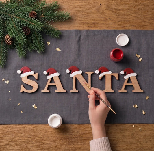 Top view of hand painting red Santa hats on laser cut plywood letters spelling SANTA on grey fabric with greenery