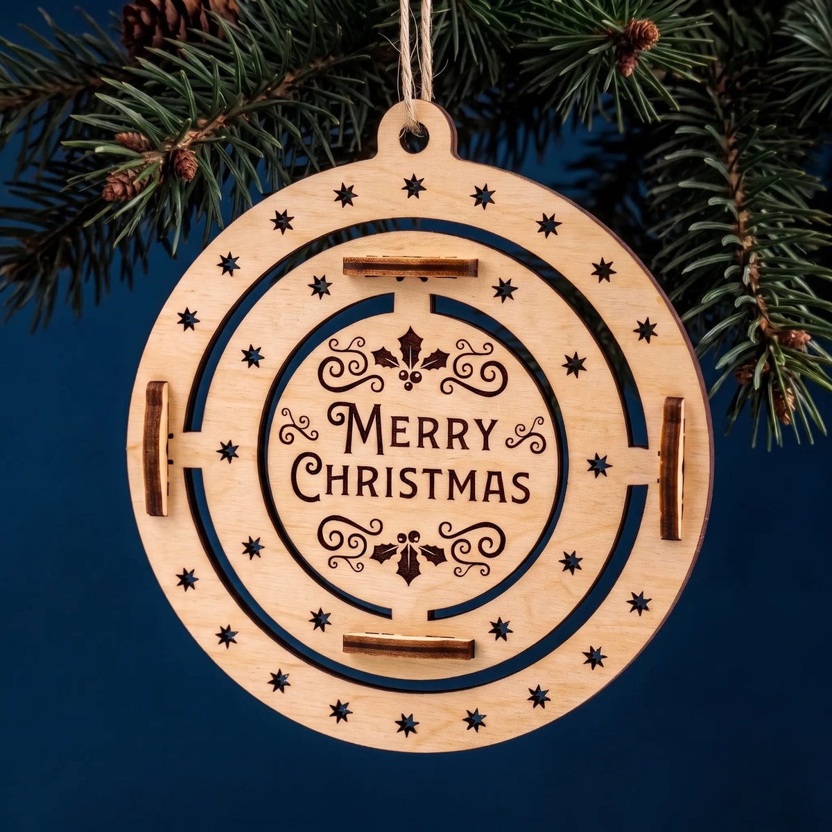 Flat laser cut plywood Merry Christmas ornament with engraved holiday flourishes hanging from pine branch on blue.