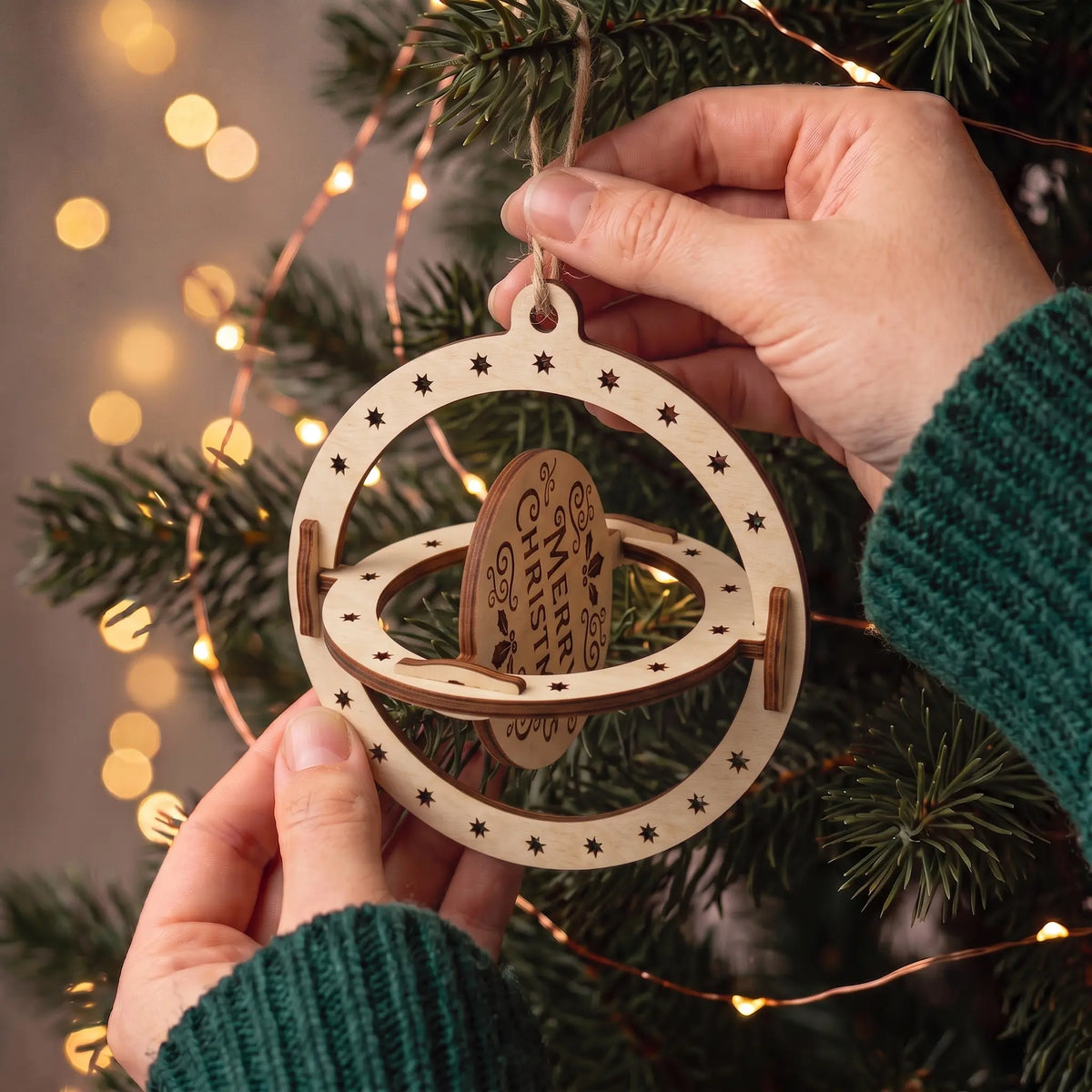 Hands adjust spinning inner rings of a laser cut plywood Merry Christmas ball ornament on lit festive tree boughs.