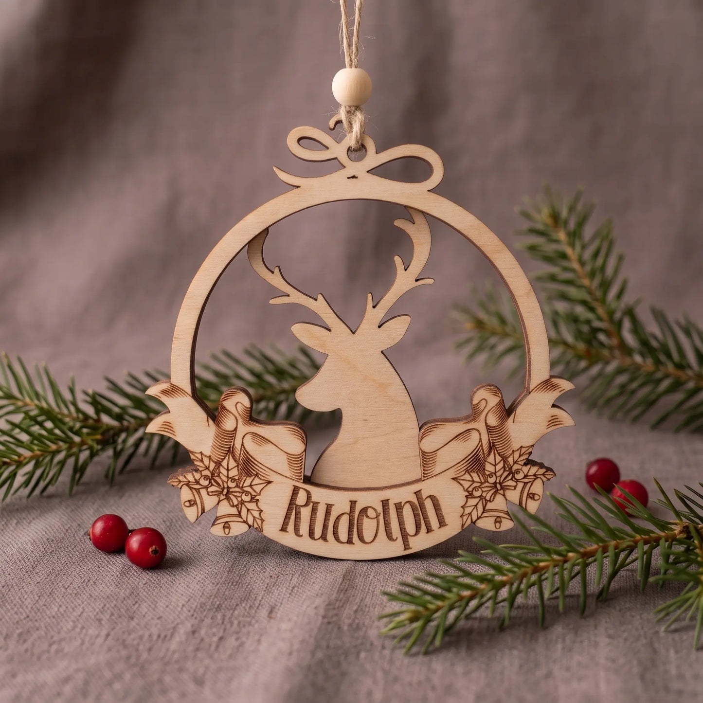 Laser cut plywood reindeer bauble with engraved Rudolph banner and bow, set on linen cloth with pine and berries.