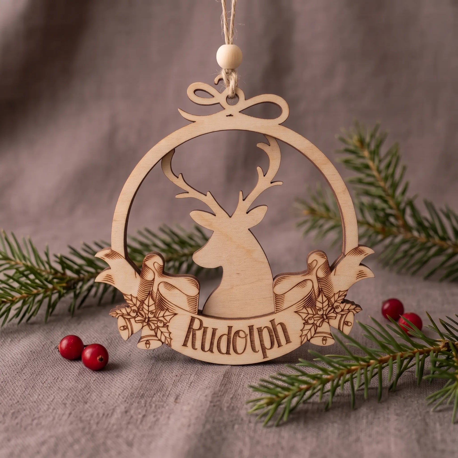 Laser cut plywood reindeer bauble with engraved Rudolph banner and bow, set on linen cloth with pine and berries.