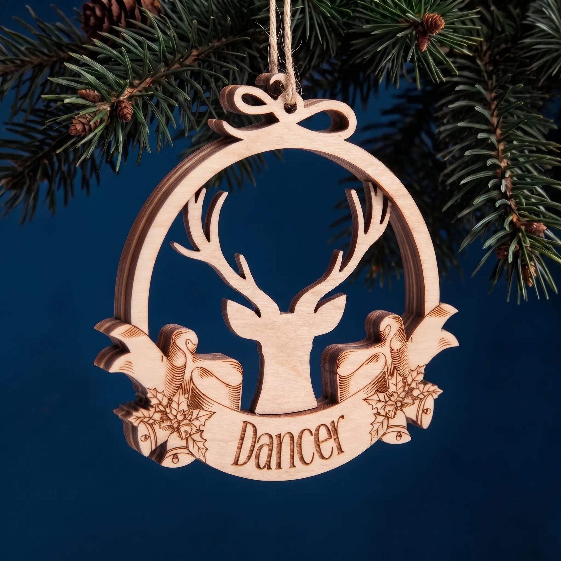 Layered laser cut plywood Dancer reindeer bauble hanging from evergreen pine needles against dark blue background.
