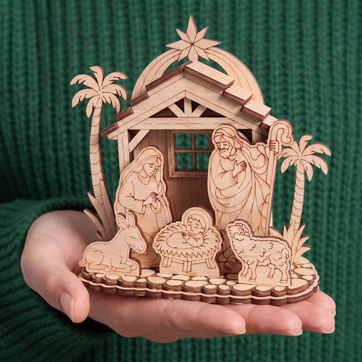 Hand holds assembled laser cut plywood nativity set with holy family and palms, showing scale against green sweater