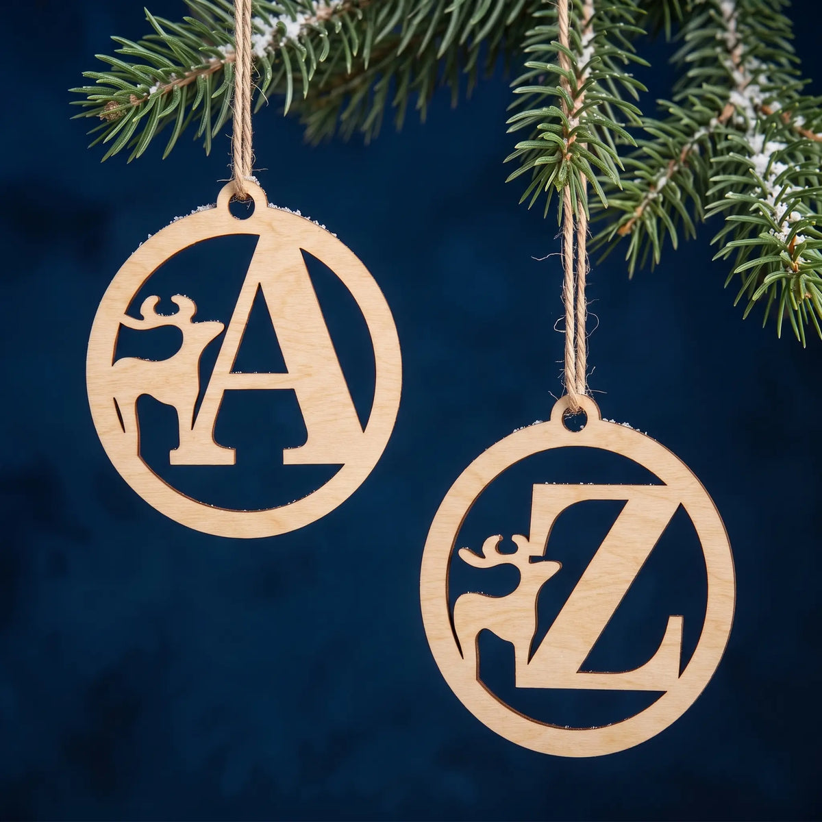 Laser cut plywood alphabet baubles with deer and letters A and Z hanging from snowy pine branch on dark blue backdrop.