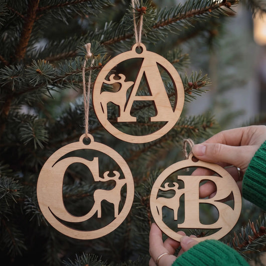 Hands display laser cut plywood letter baubles A, B, and C with deer motifs hanging on pine branches with lights