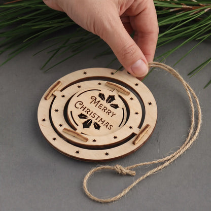 Hand holds hanging cord of flat laser cut plywood Merry Christmas disc ornament laid on grey background with pine.