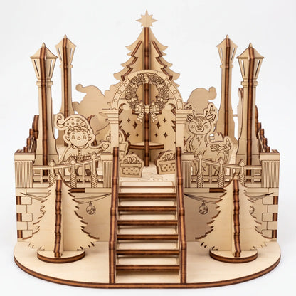 Front view of laser cut plywood holiday town square diorama with staircase, central tree, elf, and deer on white.
