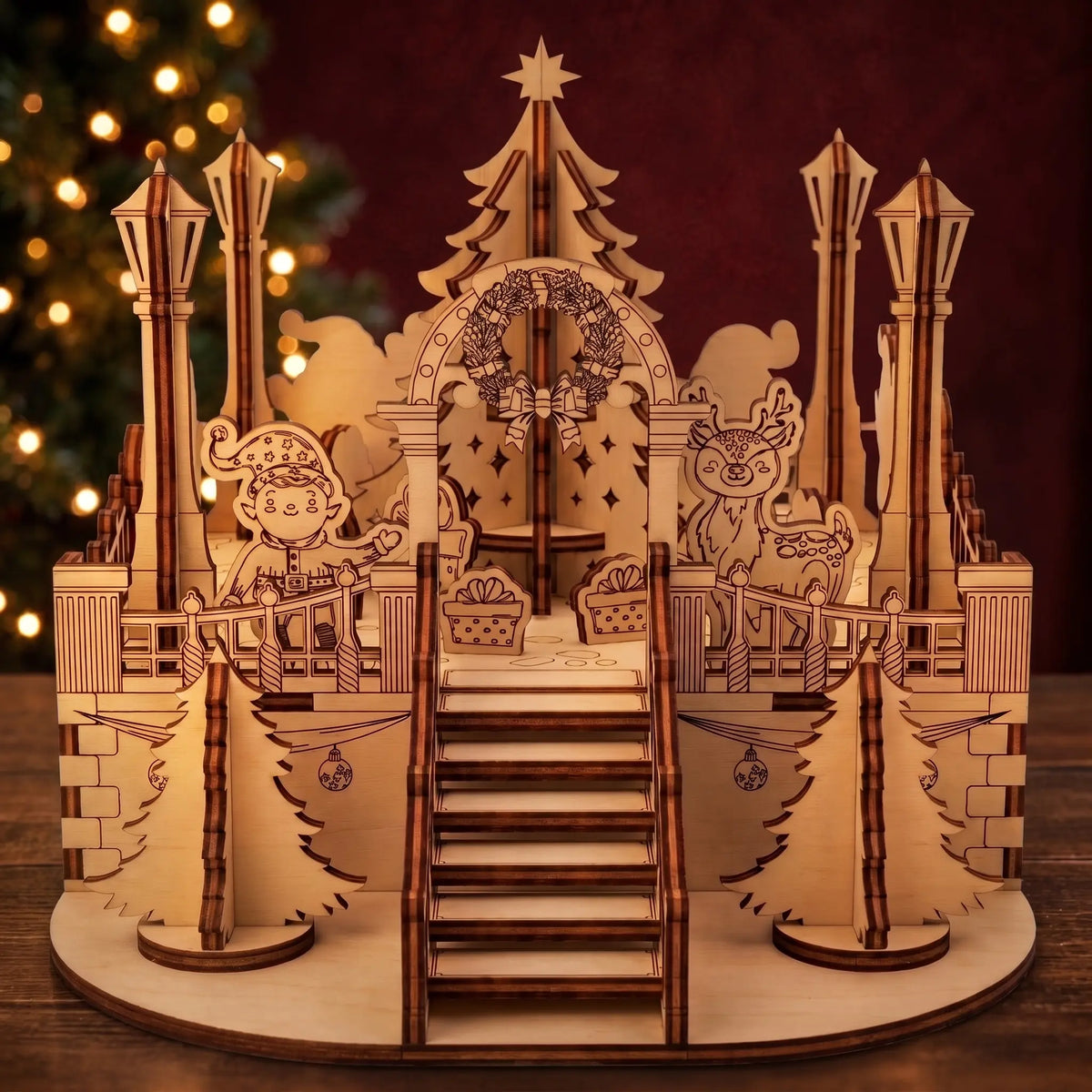 Laser cut plywood Christmas town square model with staircase, wreath arch, and elf beside glowing tree lights.