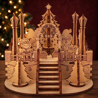 Laser cut plywood Christmas town square model with staircase, wreath arch, and elf beside glowing tree lights.