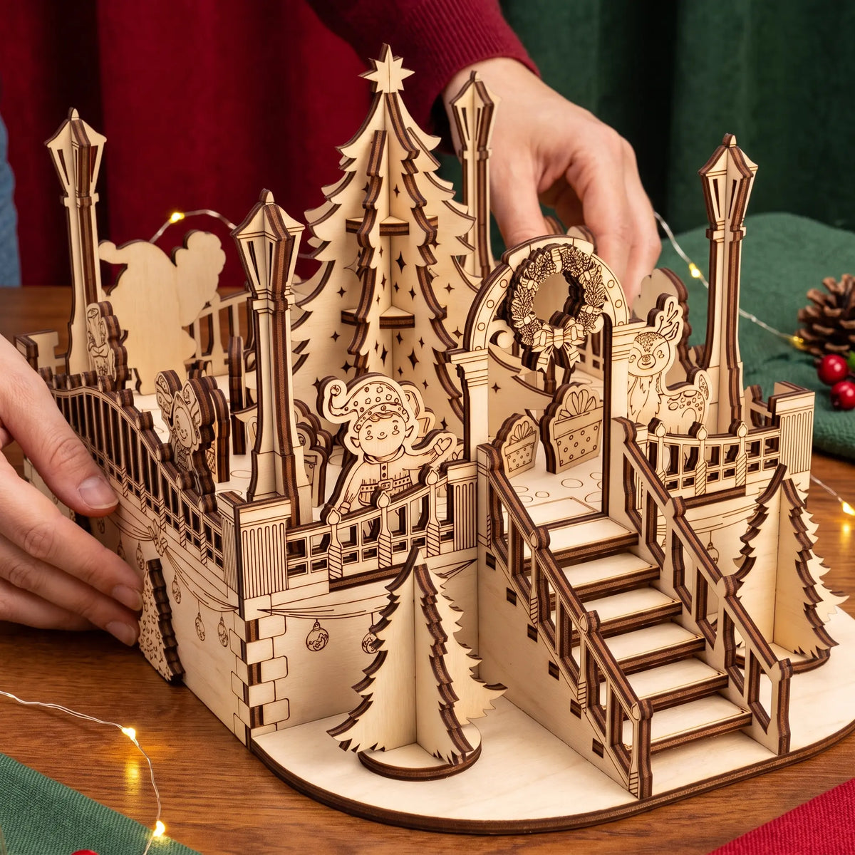 Hands position miniature laser cut plywood holiday town square diorama with lanterns, tree, and festive balcony.
