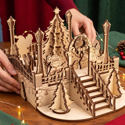 Hands position miniature laser cut plywood holiday town square diorama with lanterns, tree, and festive balcony.