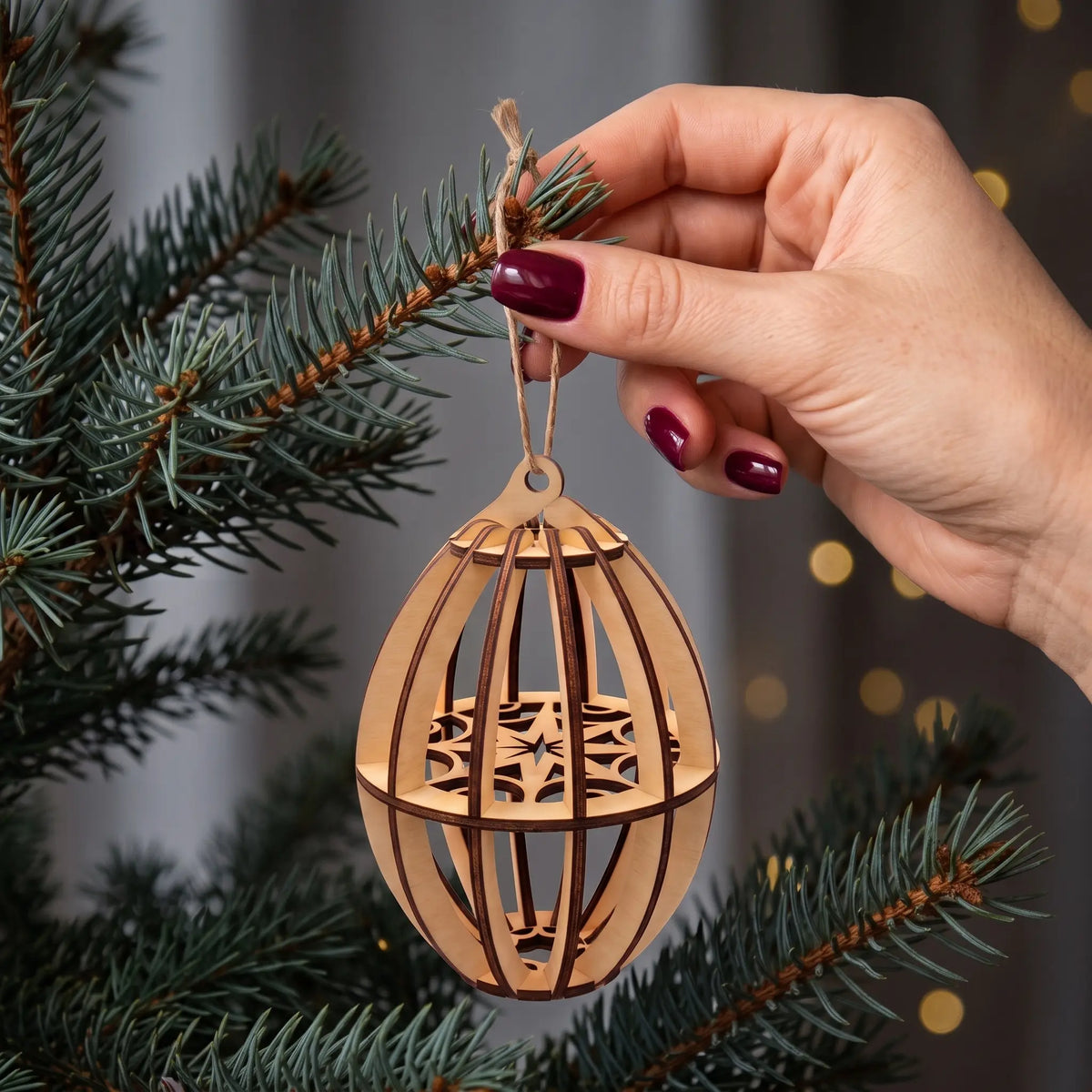 Hand hangs 3D slotted laser cut plywood oval tree ornament with snowflake center onto evergreen branches.