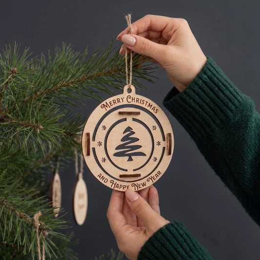 Hands hold hanging laser cut plywood Merry Christmas tree bauble, showing scale in front of pine branches