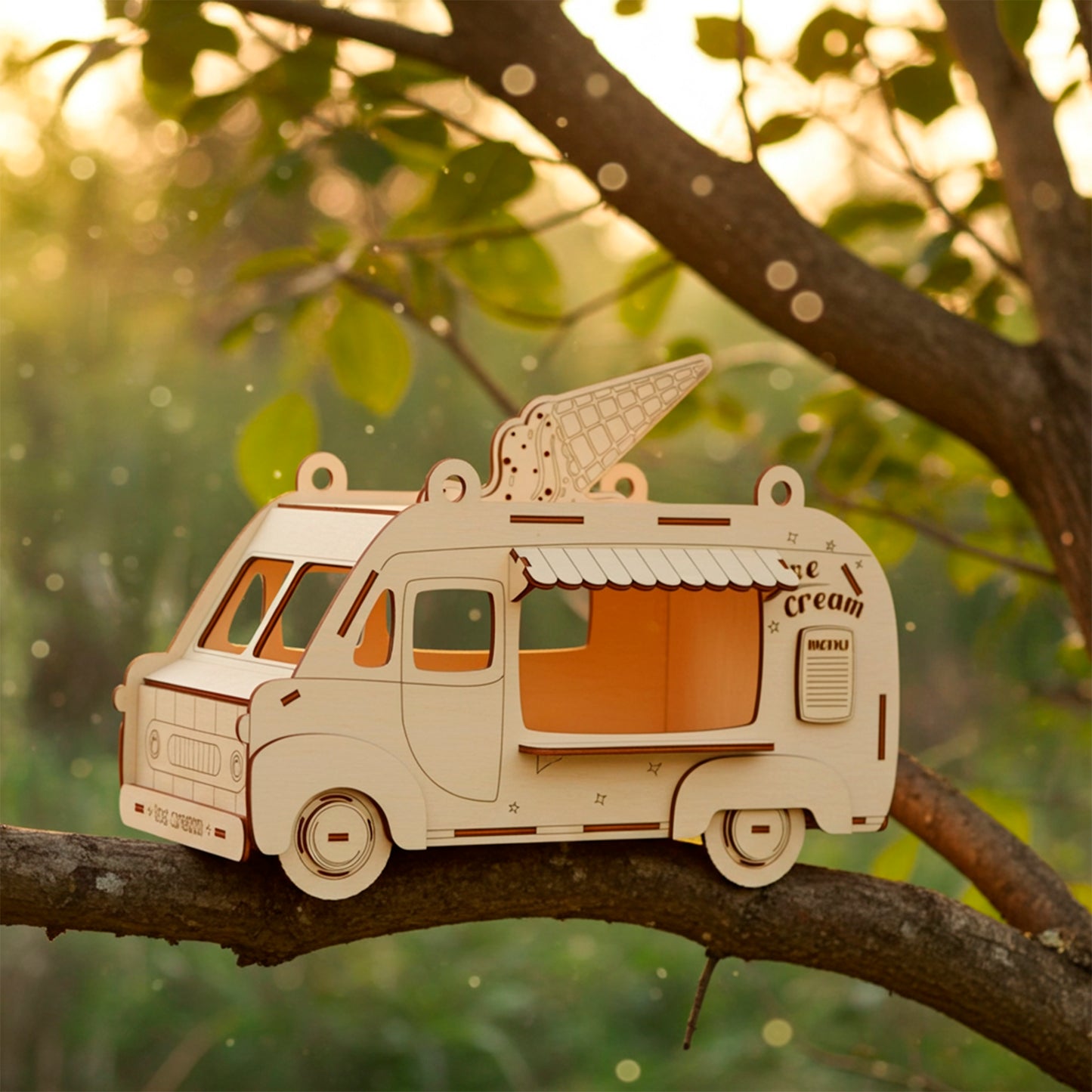 Laser cut wooden plywood Ice Cream Van Birdhouse or treat dispenser, shown lying on a green tree branch in a natural outdoor setting with warm golden hour sunlight. The model features an engraved ice cream cone on the roof, a menu sign, and wheels, showcasing the detailed DIY craft kit or ready-made whimsical gift.