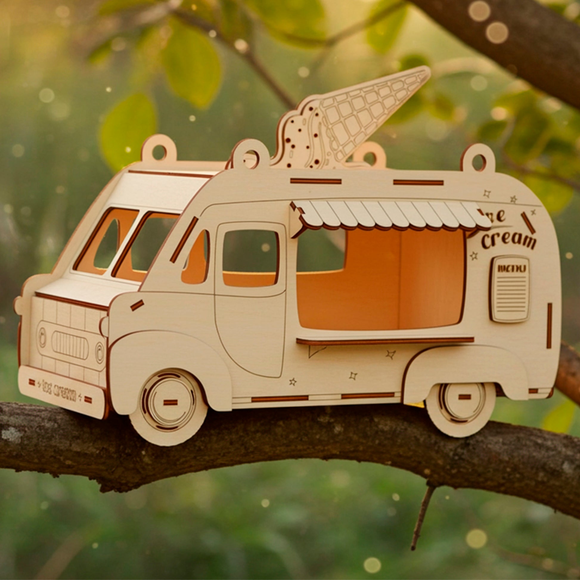 Laser cut wooden plywood Ice Cream Van Birdhouse or treat dispenser, shown lying on a green tree branch in a natural outdoor setting with warm golden hour sunlight. The model features an engraved ice cream cone on the roof, a menu sign, and wheels, showcasing the detailed DIY craft kit or ready-made whimsical gift.