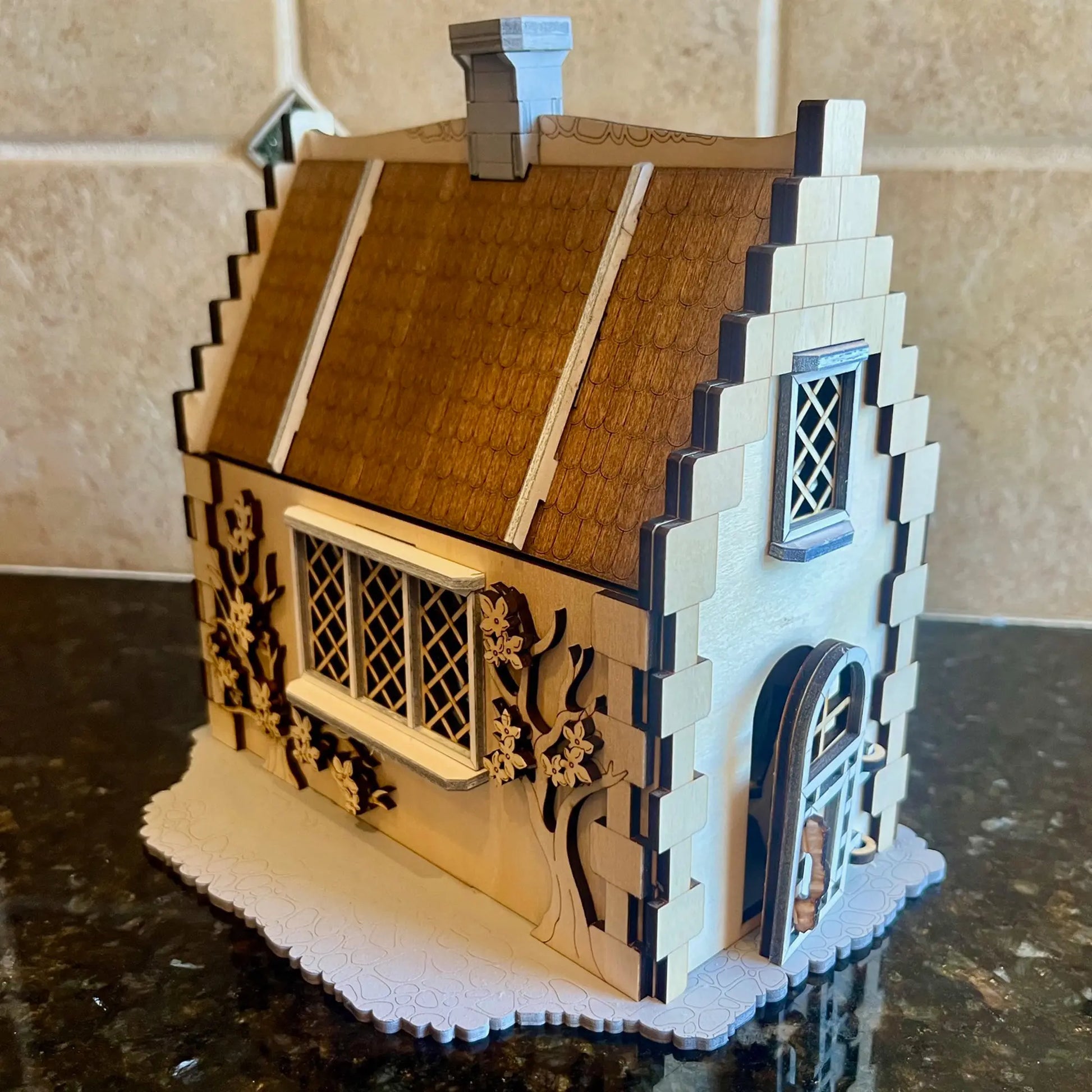 Laser cut plywood miniature cottage with a brown roof, brick chimney, and a latticed window on a reflective dark counter.