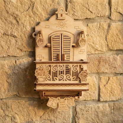 Front-facing view of a 3D laser cut plywood wall decoration shaped like a traditional European balcony. The model is mounted on a rustic stone wall, highlighting the intricate floral engravings and layered wooden construction.