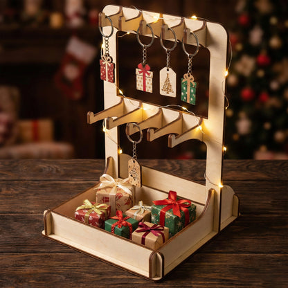 Two-tier laser cut wooden plywood  stand decorated with fairy lights for a holiday market. The display features Christmas-themed keychains shaped like presents and trees, with small wrapped gift boxes in the bottom tray against a blurred Christmas tree background.