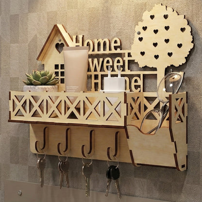 Wooden laser cut keyholder "Home Sweet Home" with a charming house silhouette, lattice balcony shelf for small items, multiple hooks for keys, and a heart-filled tree detail, mounted on a textured wall background.