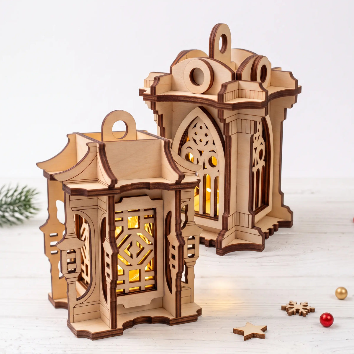 Pair of glowing laser cut plywood holiday lanterns standing on white painted wooden surface with small festive decor.