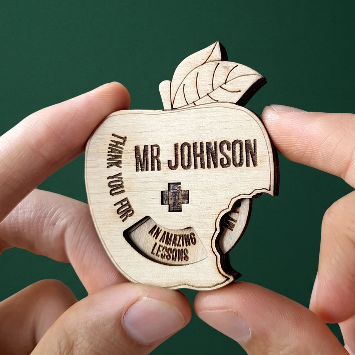 Hands holding personalized laser cut plywood apple teacher appreciation spinner token against solid dark green background.