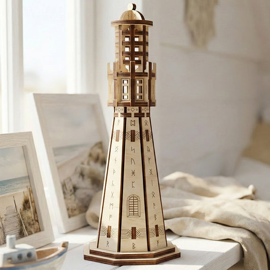 A tall, detailed laser cut plywood lighthouse standing on a white shelf. The wooden model shows dark burnt edges and engraved runic symbols. Two framed beach photos and a soft linen cloth are in the background, showing the item's scale.